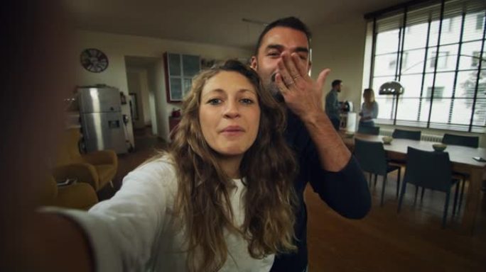 年轻的新婚夫妇的真实照片正在家里的厨房里用智能手机给亲戚或朋友自拍或视频通话。