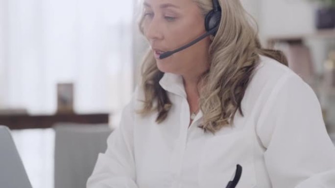 一个成熟的呼叫中心代理在使用耳机和笔记本电脑帮助并与她的家庭办公室中的客户交谈时写作。远程自由职业者