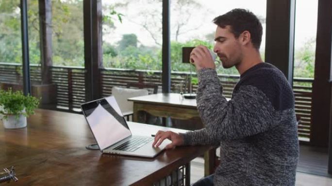 白人男子在家工作，使用笔记本电脑并在饭厅喝咖啡
