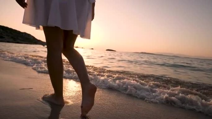 一个穿着白色太阳裙的年轻无忧无虑的女人在日落度假旅行期间沿着海滨沙滩散步的真实特写镜头。