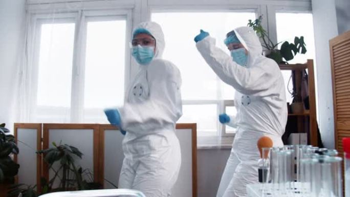两位快乐有趣的女科学家，穿着白色医疗防护服的医生跳舞，庆祝实验室研究的成功。