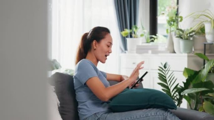 年轻的亚洲女性穿休闲网站在沙发上使用智能手机感觉令人兴奋的滚动屏幕在线购物在客厅室内植物在家里。