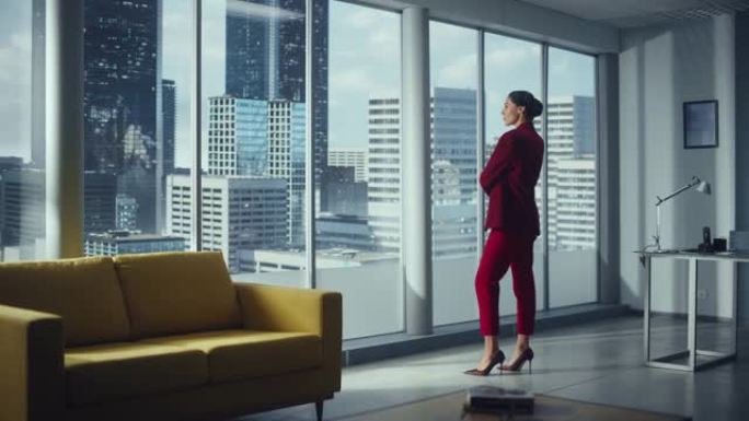 成功的体贴女商人穿着完美的红色西装站在办公室，看着窗外的大城市。自信的女性公司IT公司最高经理计划投