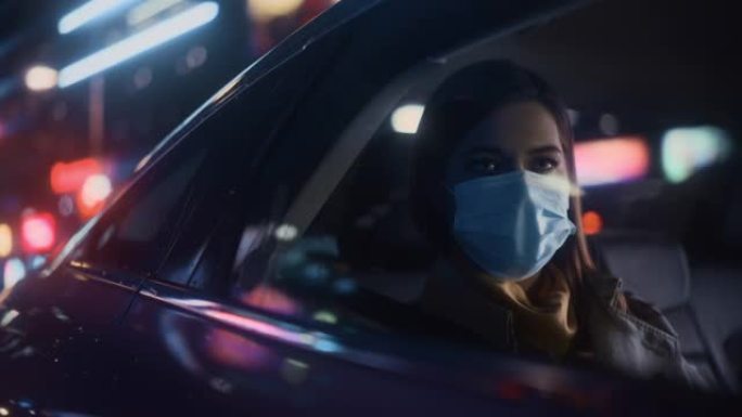 戴着口罩的女性晚上在出租车的后座上通勤回家。美丽的女乘客在城市街道上开着霓虹灯的汽车时看着窗外。