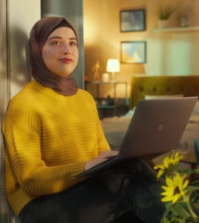 垂直屏幕: 戴着头巾的富有创造力的穆斯林女性坐在舒适公寓的窗台上，在笔记本电脑上工作。公司专家在家远