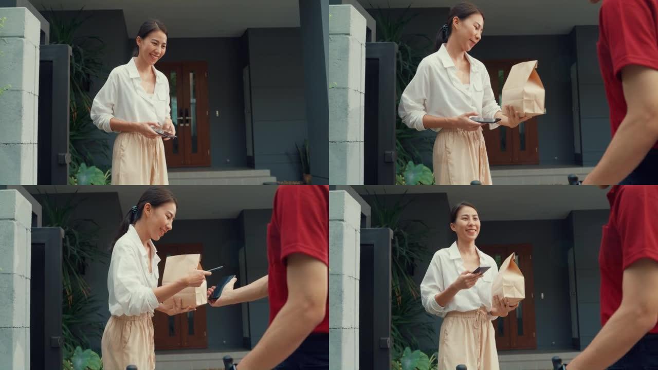 年轻快乐的亚洲女人接受送货员送的纸袋，带红色制服的自行车携带箱在房子前面的智能手机上扫描二维码付款。