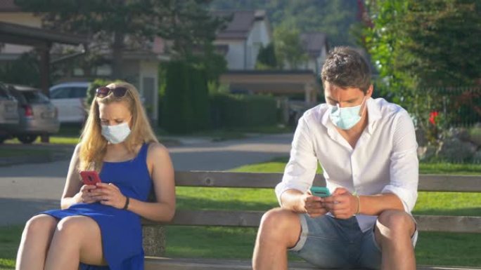 特写: 两个陌生人坐在公园长椅上发短信时戴着口罩。