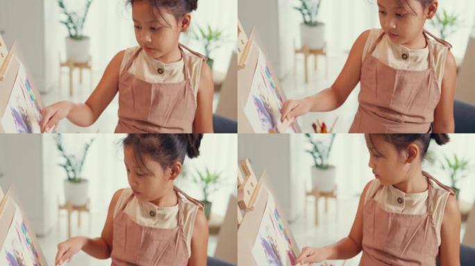 特写带围裙的亚洲学步女孩坐在沙发椅上，在带调色板的油画画布前使用画笔专注于周末在家绘画抽象。