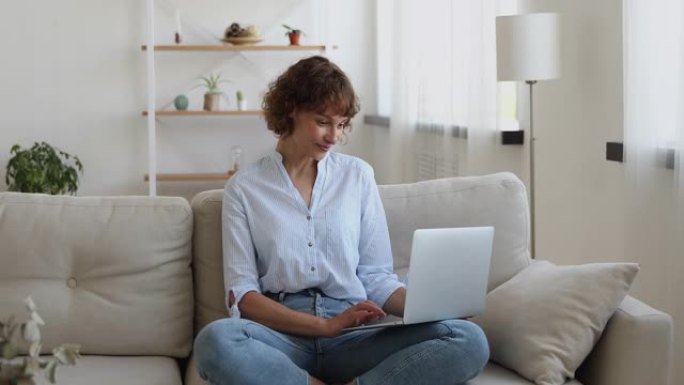 放松的40多岁的女人坐在沙发上盘腿使用笔记本电脑