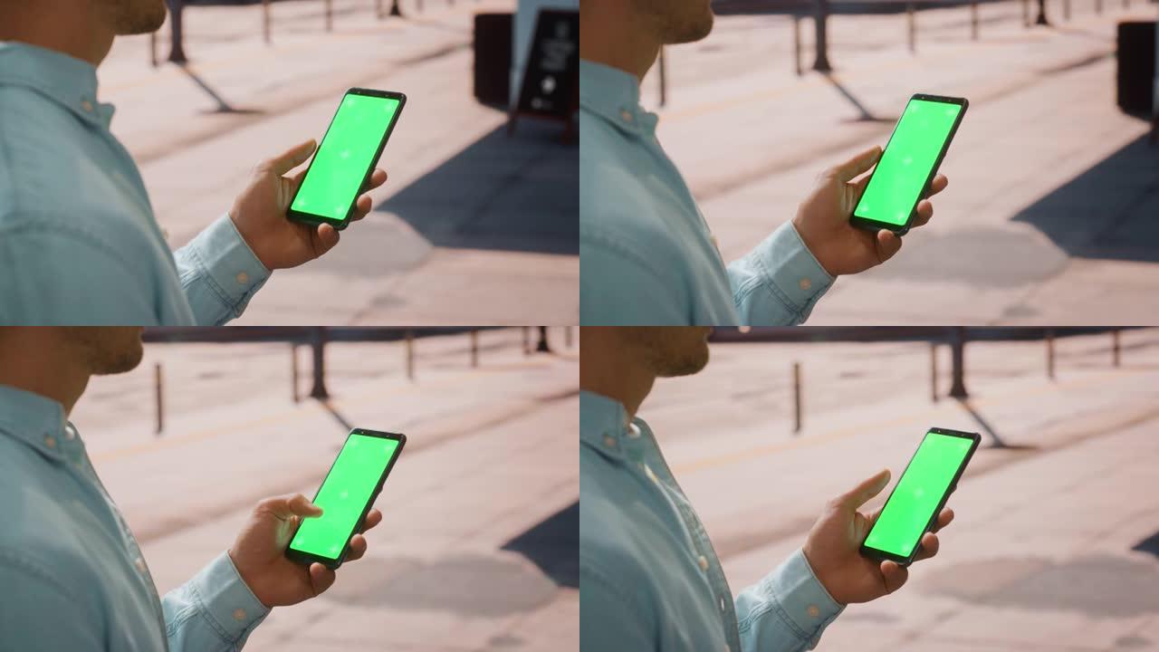 在阳光明媚的日子里，一个年轻人使用带有绿色屏幕色度键模拟显示的智能手机在街道上垂直显示。他正在轻敲屏