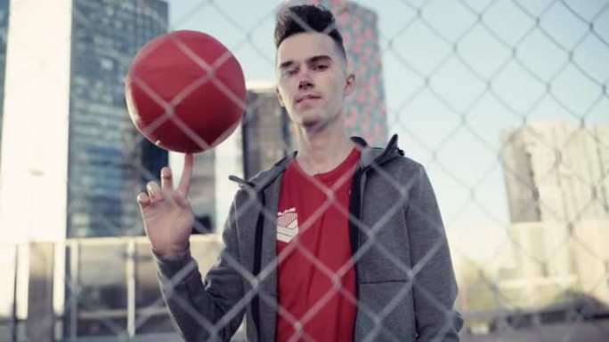 一位时尚的年轻足球运动员在镜头前摆姿势，手指上旋转一个红球的肖像。英俊的足球运动员站在城市的篱笆后面
