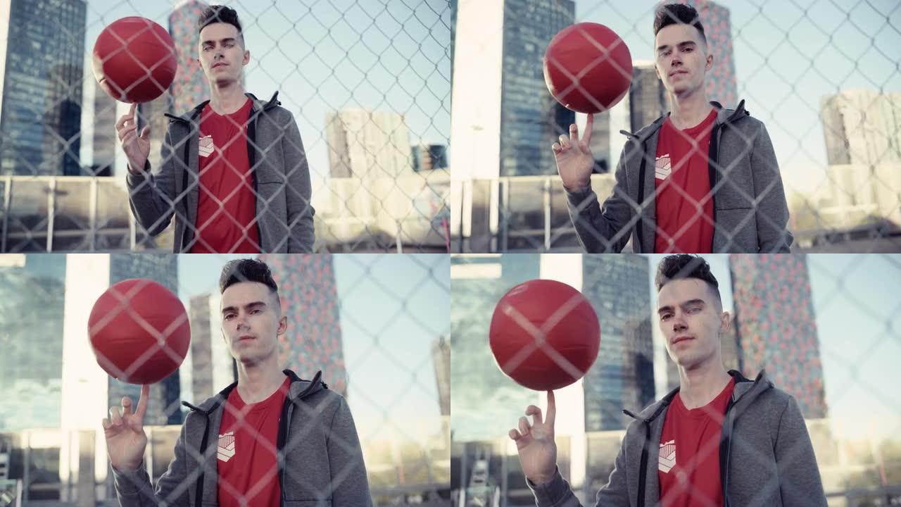 一位时尚的年轻足球运动员在镜头前摆姿势，手指上旋转一个红球的肖像。英俊的足球运动员站在城市的篱笆后面