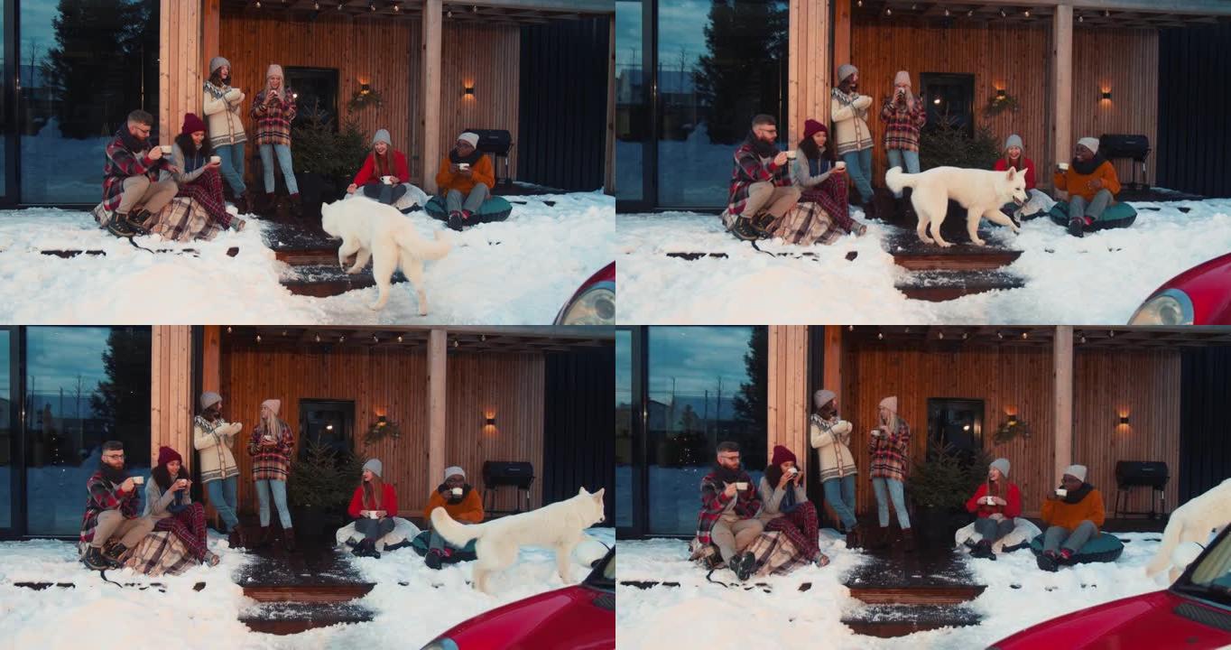 一群快乐放松的多民族朋友在舒适的寒冷冬季房子露台上喝热茶，带狗，红色汽车慢动作