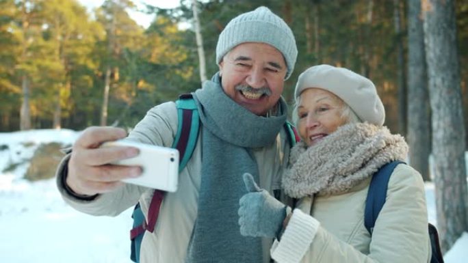 冬日在户外自拍的老年夫妇微笑着用智能手机摄像头竖起大拇指