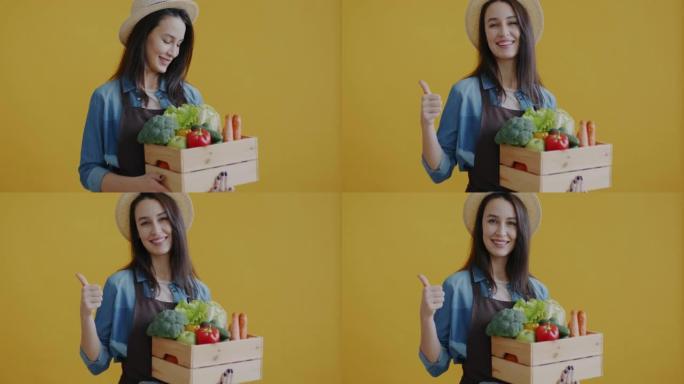 欢快的园丁的肖像拿着新鲜蔬菜的木箱，在黄色背景上显示竖起大拇指的手势