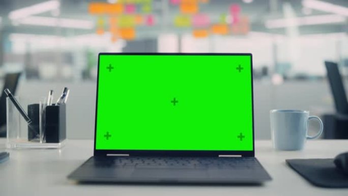 带有模拟绿屏色度键显示屏的笔记本电脑站在现代创意办公室的桌子上。背景玻璃墙上有五颜六色的规划笔记。放