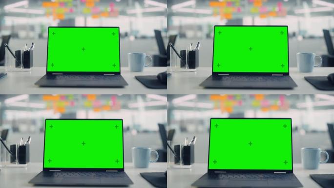 带有模拟绿屏色度键显示屏的笔记本电脑站在现代创意办公室的桌子上。背景玻璃墙上有五颜六色的规划笔记。放