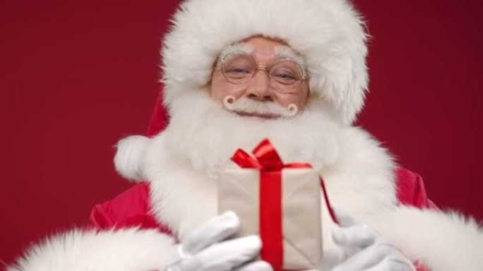 一位善良的圣诞老人站在红色的背景下，微笑着在相机中展示了一份可爱的小礼物，以至于只有礼物在焦点上，然