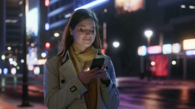 一个穿着风衣的美女的肖像在夜晚霓虹灯的现代城市街道上行走。迷人的女性使用智能手机，环顾城市电影环境。