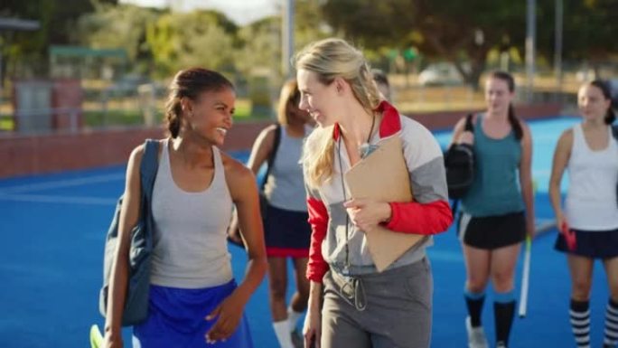 训练后，顽皮的女子曲棍球教练与一名年轻女子球员交谈。曲棍球运动员与体育老师一起散步，在运动场上进行了