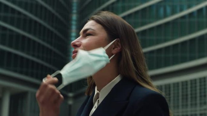 年轻的女商人脱下医用防护口罩，在摩天大楼的背景下深呼吸的电影特写镜头。企业概念，保护，病毒传输，安全