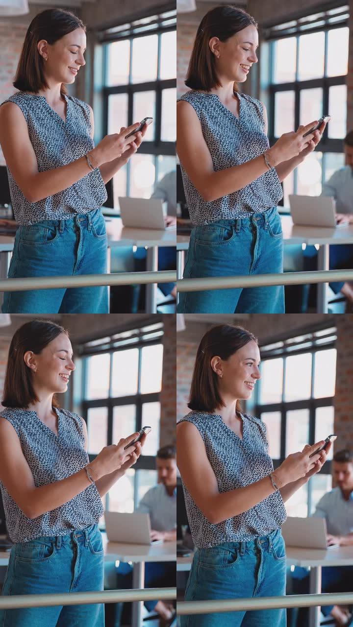 手机上微笑的女商人站在繁忙的办公室留言的垂直视频