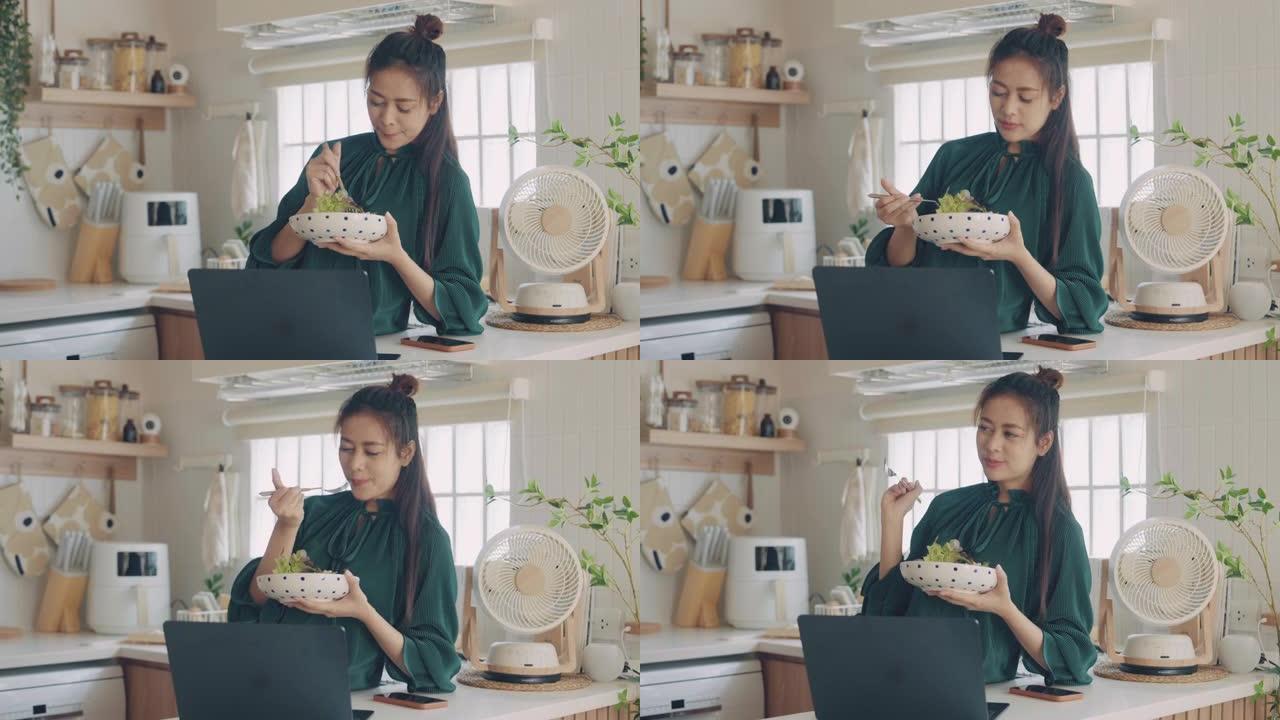 美丽的年轻亚洲妇女在家里吃健康的食物，在厨房使用笔记本电脑。