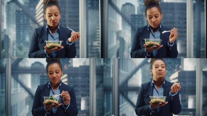 自信的黑人女性乘坐玻璃电梯到现代商务中心的办公室，吃健康的蔬菜沙拉。快乐的经理为工作中富有成效的一天