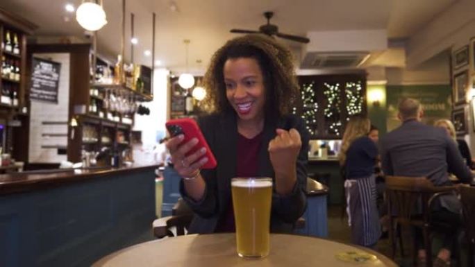 成功的女商人在酒吧喝啤酒的同时，在收到消息后检查智能手机并庆祝。
