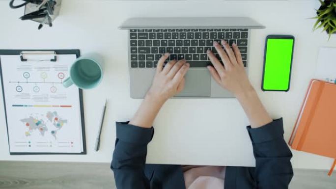女性双手在办公室与笔记本电脑一起工作的俯视图，而绿色色度键屏幕智能手机在桌子上可见