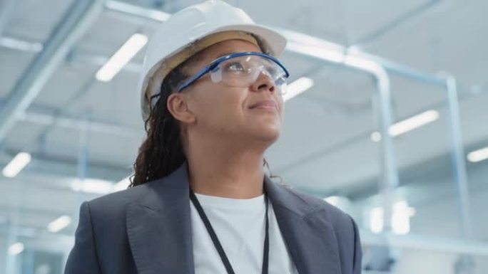 站在电子制造工厂的一位成功，快乐的中年非洲女工程师的特写肖像，戴着白色安全帽。黑人重工业专家摆姿势拍