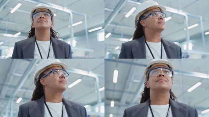 站在电子制造工厂的一位成功，快乐的中年非洲女工程师的特写肖像，戴着白色安全帽。黑人重工业专家摆姿势拍