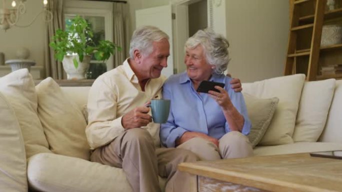 白人高级夫妇一起坐在家里的沙发上，一边微笑着使用智能手机