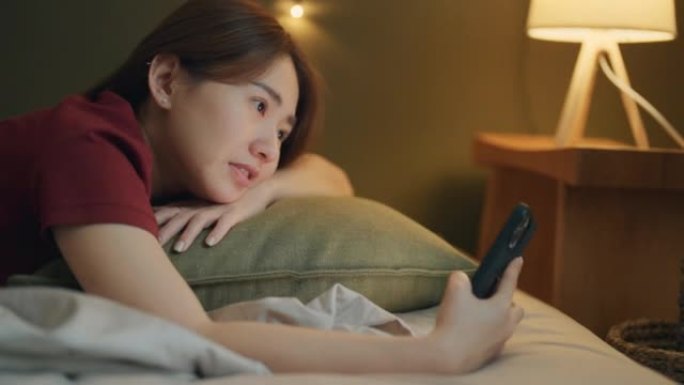 在酒店舒适的床上穿着休闲服并使用智能手机的快乐亚洲女性
