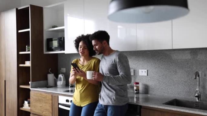 幸福的夫妇靠在厨房的柜台上，一边说话一边微笑着看着智能手机上的东西