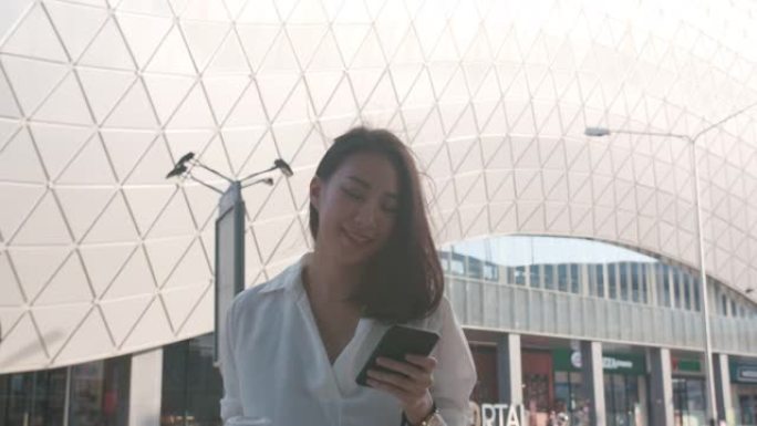 亚洲女商人穿着时尚的办公室衣服，拿着一次性纸杯热饮，在城市现代城户外散步时使用智能手机。