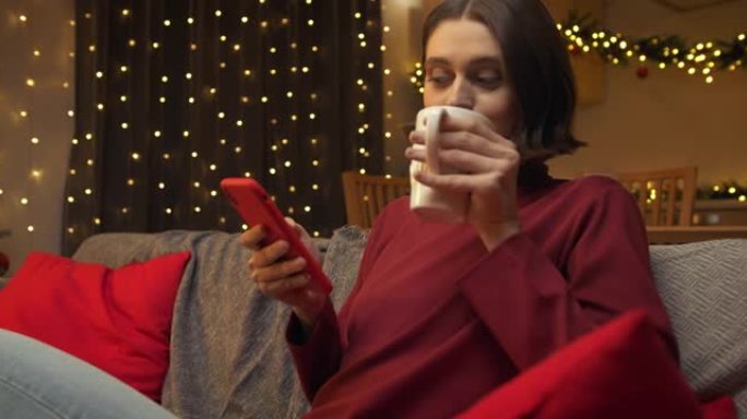 一位穿着红色毛衣的可爱年轻女子正在她的红色智能手机上发短信，坐在圣诞节装饰的房间的沙发上，端着一杯茶