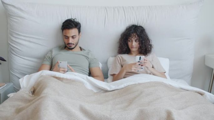 混血夫妇躺在床上并在休闲时间使用智能手机的肖像
