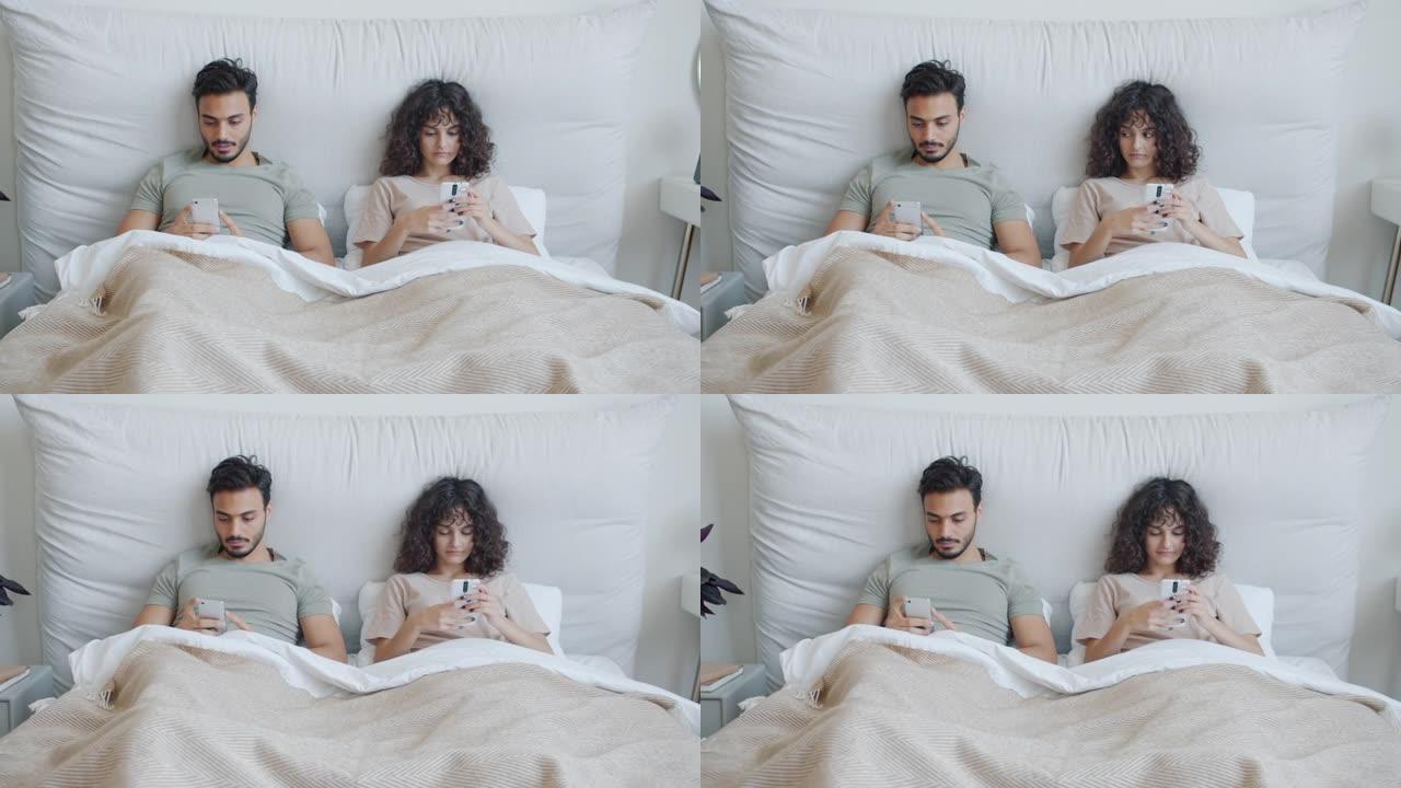 混血夫妇躺在床上并在休闲时间使用智能手机的肖像