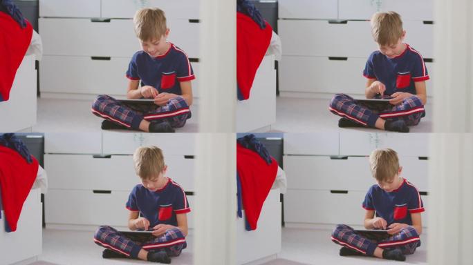 男孩在卧室里穿着睡衣坐在地板上的数字平板电脑上玩耍