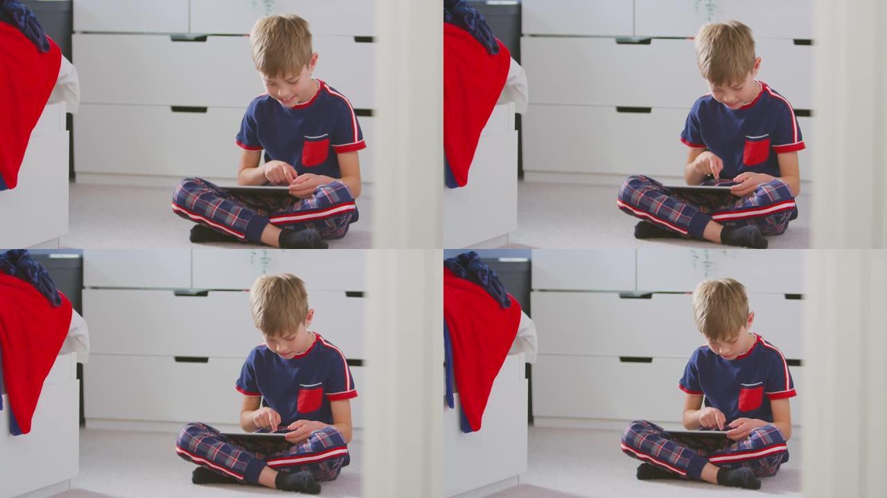 男孩在卧室里穿着睡衣坐在地板上的数字平板电脑上玩耍