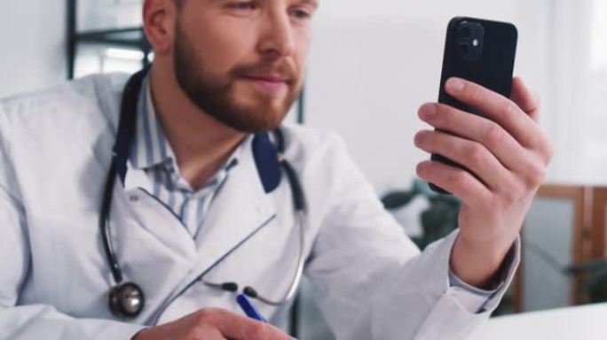 特写年轻的白人男性医生使用智能手机网络会议咨询远程患者，在办公室做笔记。