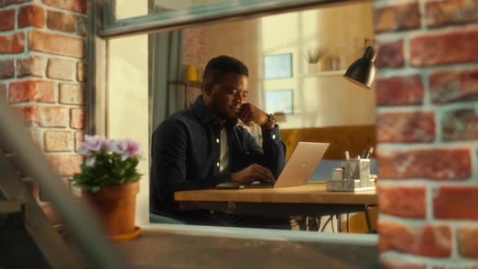 年轻英俊的男性使用笔记本电脑，坐在公寓的客厅。非裔美国商人回复电子邮件，在家写商业计划书。公寓窗户内
