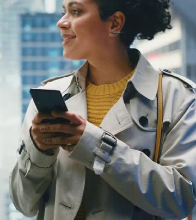 垂直屏幕: 现代商务中心的黑色女性乘坐玻璃电梯到办公室。成功的经理在使用智能手机时微笑，写短信，在电