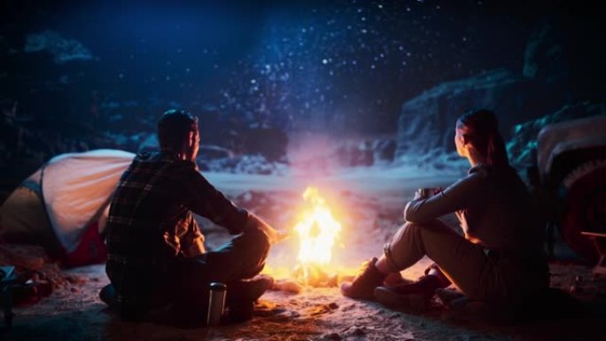 幸福的夫妻坐在篝火旁分享一杯饮料，在峡谷中露营时看着夜空。两个旅行的年轻人讲述有关生活的励志故事，看