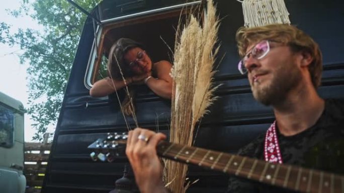 年轻的嬉皮士女士弹吉他，而女友则在露营者中欣赏音乐的声音