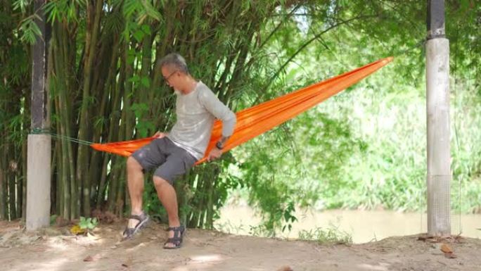 亚洲华裔高级男子在竹树下河边的吊床上躺着享受他的下午