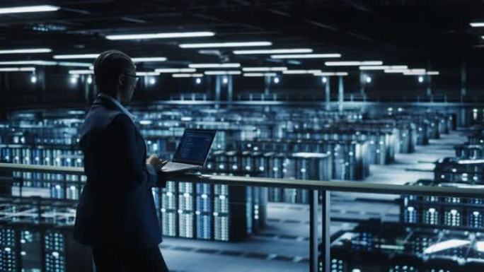 女性IT专家在数据中心使用笔记本电脑，走在俯瞰大服务器场云计算设施的桥上。女商人，电子商务企业家。基