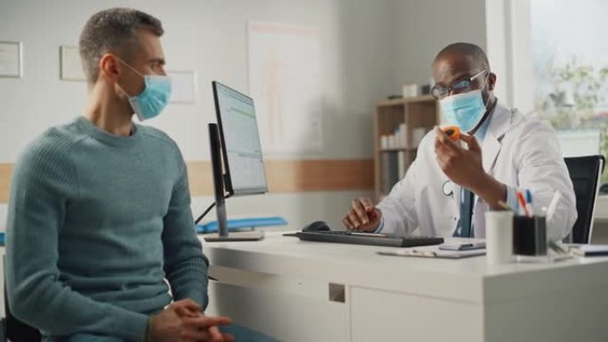 一位非裔美国家庭医生正在一家诊所与一位年轻男性病人交谈。两人都要戴口罩。穿着白大褂的医生坐在医院办公