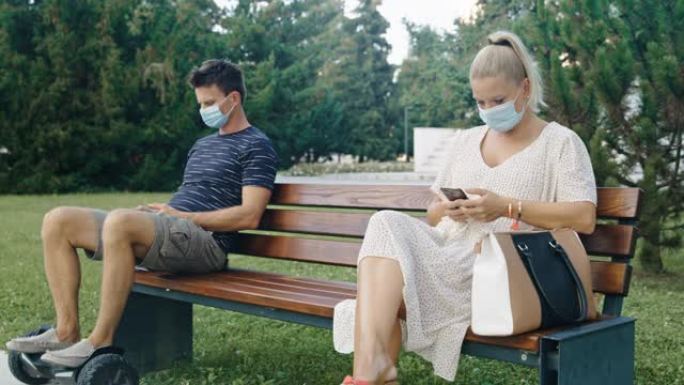 SLO MO夫妇在公共公园的长凳上使用手机戴着防护口罩